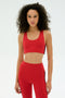 A woman with curly hair wears the SPLITS59 Ella Airweight Bra in Pirate Red/Indigo, paired with high-waisted leggings, both made from stretchy spandex, standing against a plain white background.