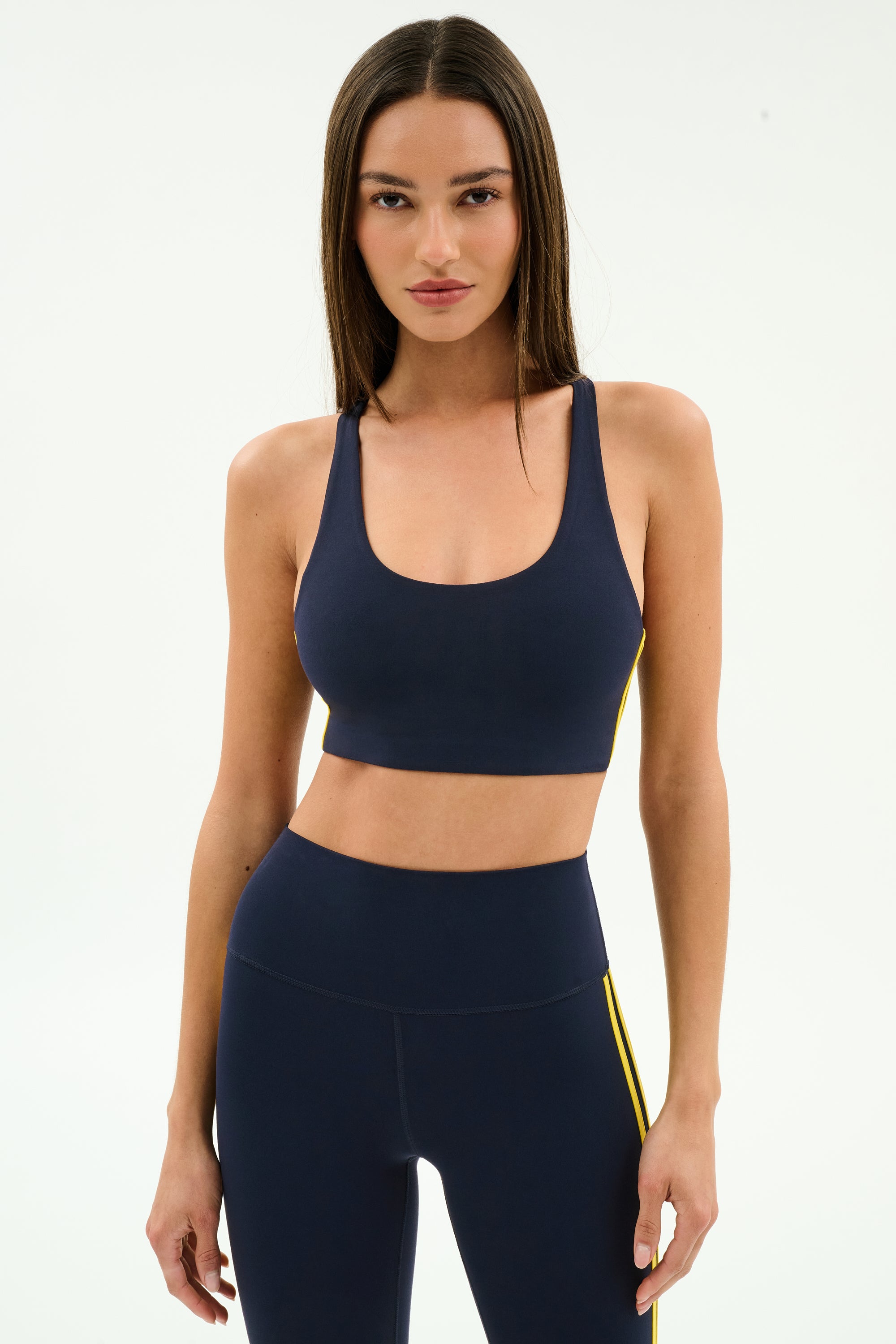 A woman wears the SPLITS59 Ella Airweight Bra in Indigo/Bright Yellow with matching high-waisted leggings, featuring yellow side stripes and stretch fabric, standing against a plain white background.