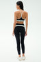 A woman faces away in a black SPLITS59 Emerson Airweight High Waist - Black/Ice legging with white accents and a black sports bra, paired with white sneakers—ideal for Pilates or hot yoga—against a plain white background.