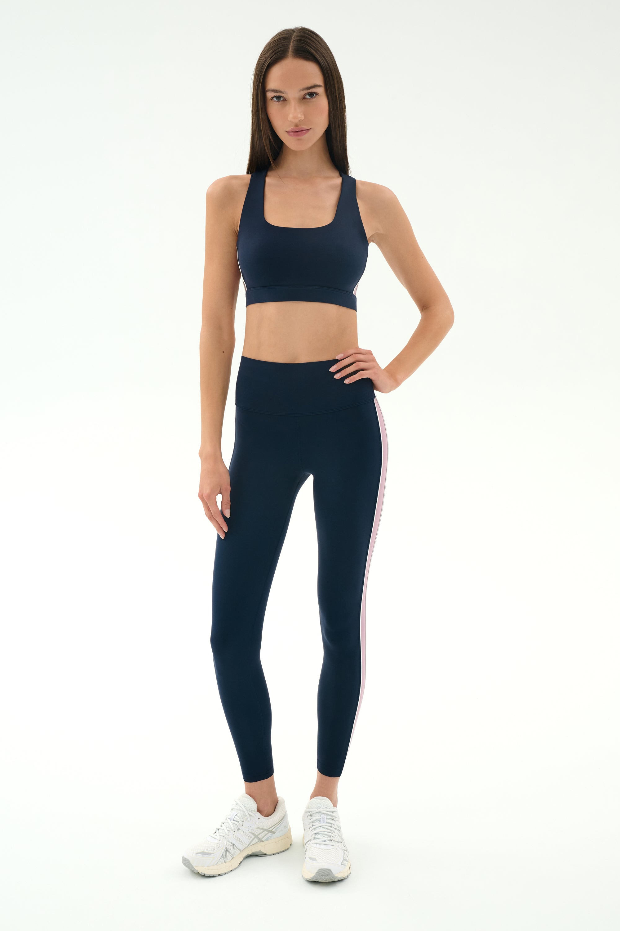 A woman stands against a white background wearing SPLITS59's Margot Rigor Legging in Indigo/Blush and a navy sports bra, paired with white athletic shoes.