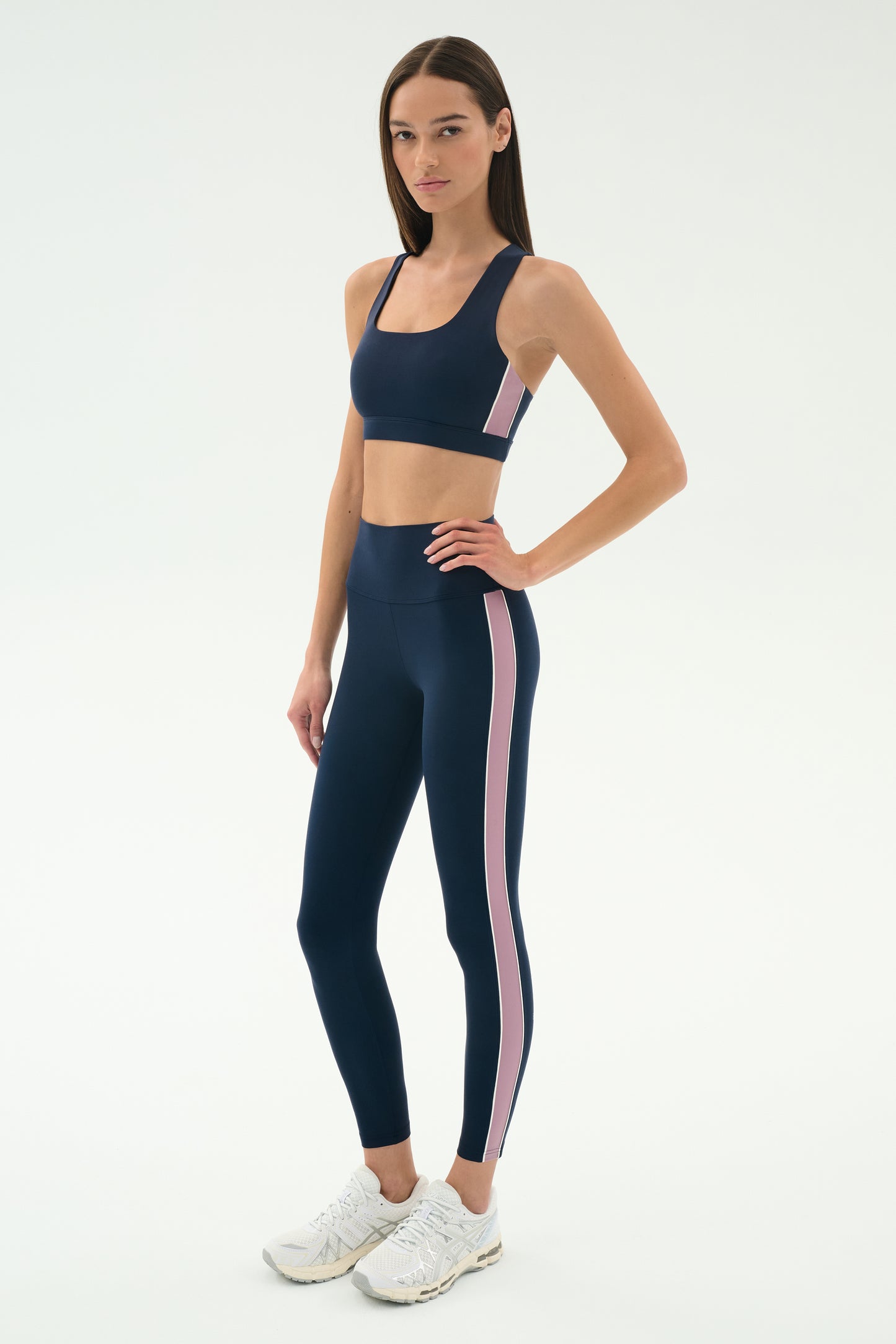 A woman stands in a studio wearing the SPLITS59 Margot Rigor Legging in Indigo/Blush, paired with a navy sports bra and white athletic shoes.
