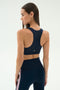 A woman is shown from the back in the SPLITS59 Lexi Rigor Bra - Indigo/Blush, a navy sports bra with matching leggings featuring pink side stripes, against a plain light background.