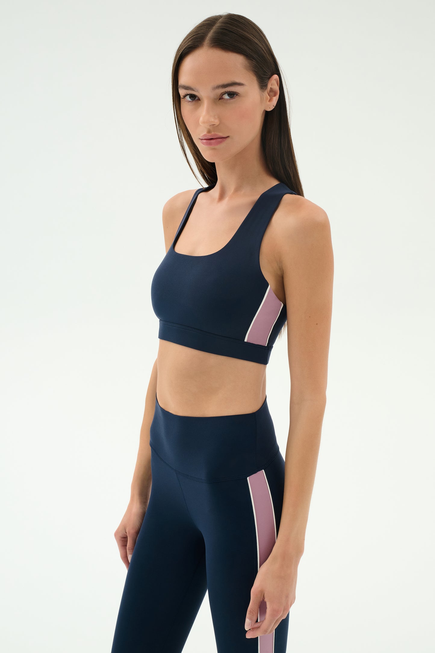 A woman wears the SPLITS59 Lexi Rigor Bra in Indigo/Blush, paired with matching high-waisted leggings featuring pink side stripes, posing against a plain light background.