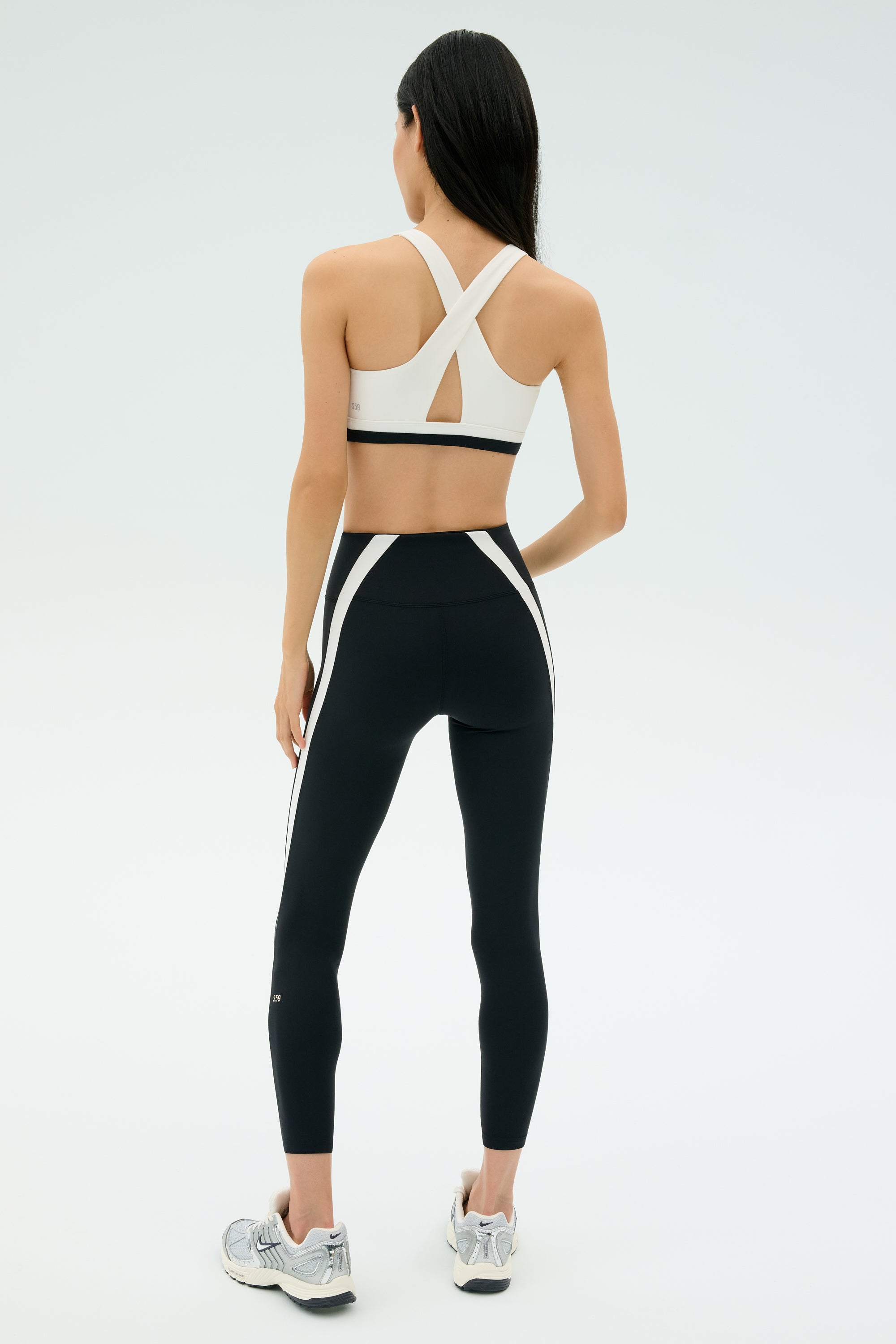 A woman faces away, wearing SPLITS59 Miles High Waist Rigor 7/8 leggings in black/white with a white sports bra and silver sneakers, ready for her workout against a plain light background.