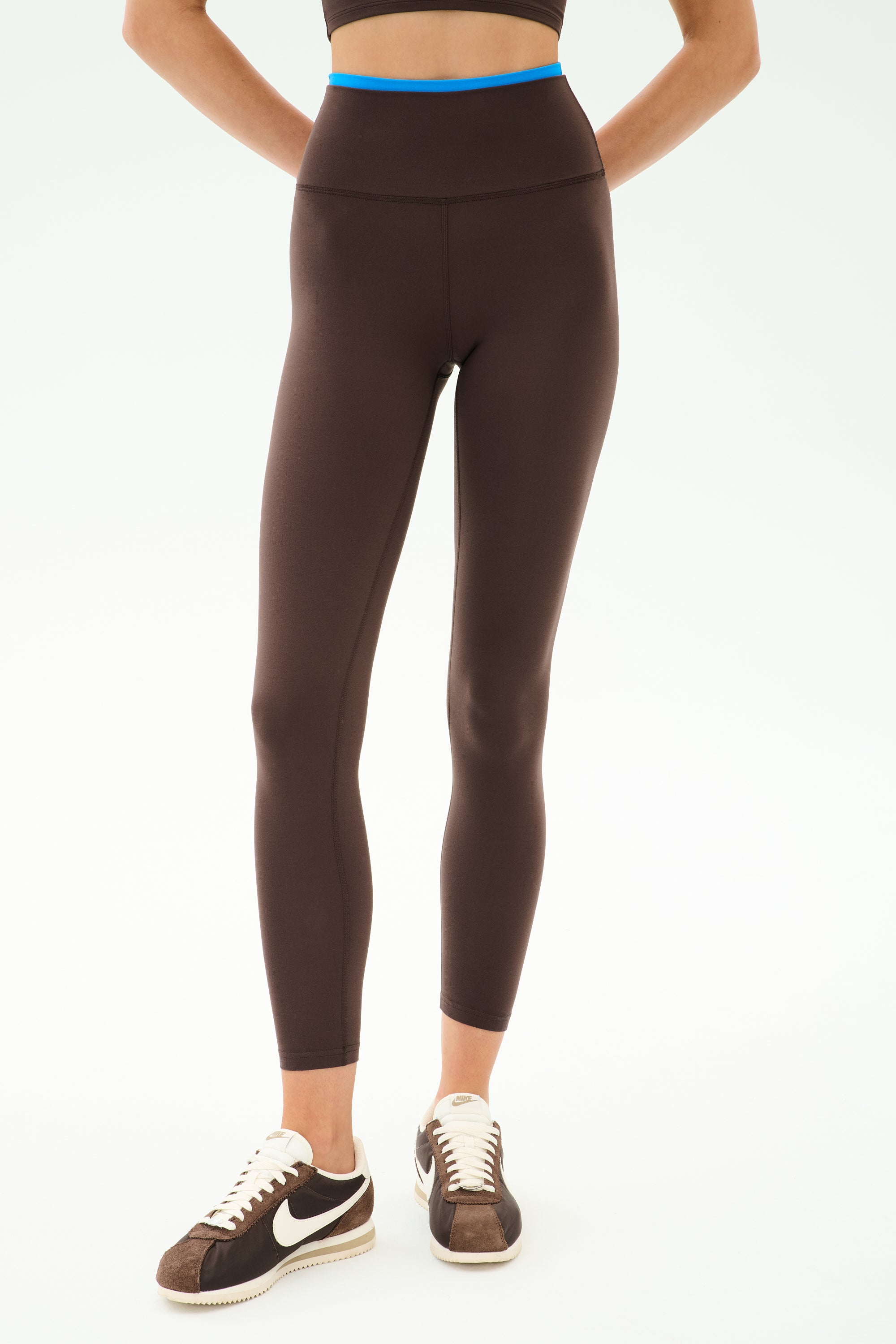 A person wears SPLITS59 Dual High Waist Airweight 7/8 leggings in Dark Chocolate/Classic Blue, paired with a black sports bra and brown and white Nike sneakers, standing against a plain light background—ideal for hot yoga.