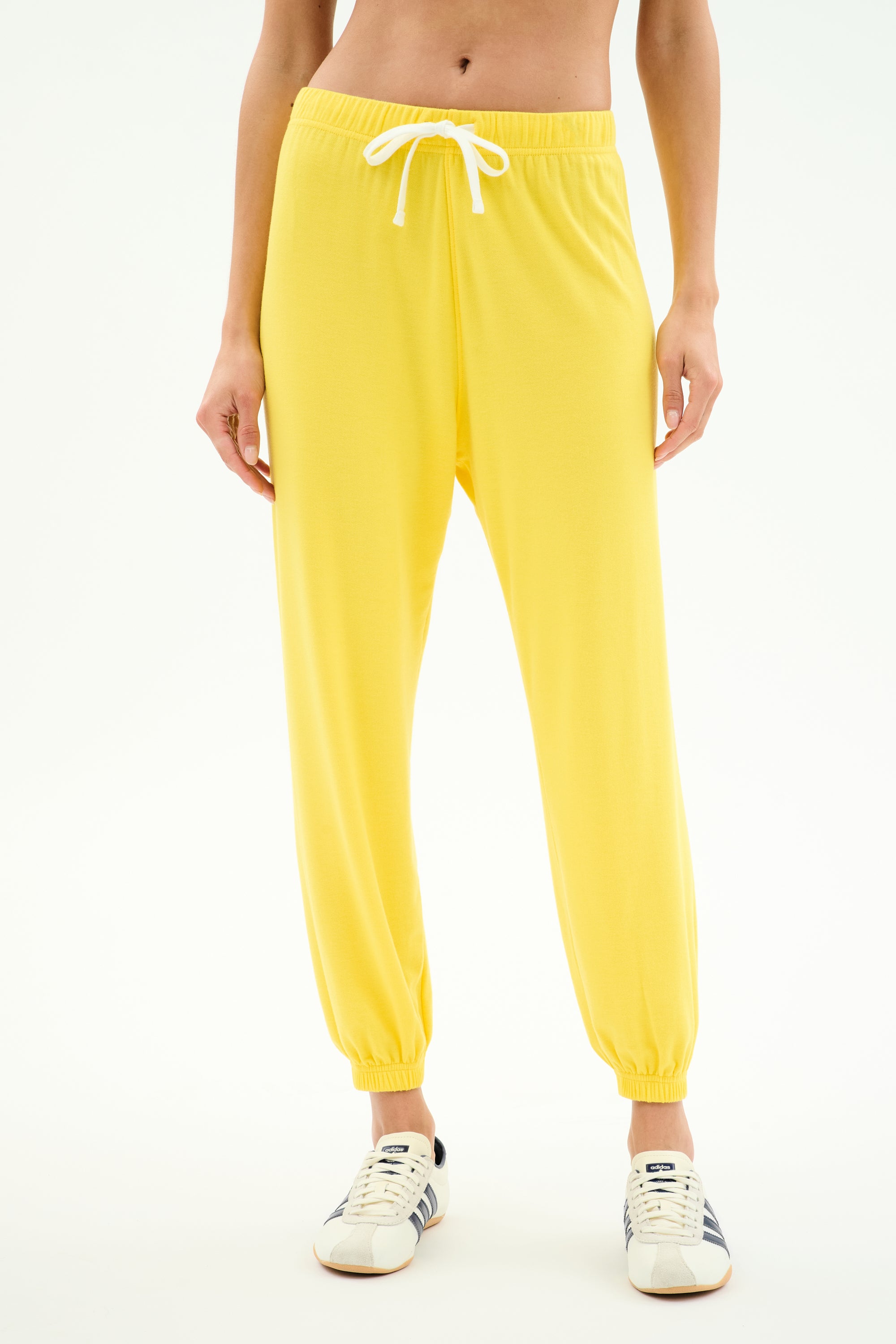 A person wears SPLITS59's Andie Oversized Fleece Sweatpant in bright yellow, featuring a white drawstring, paired with white sneakers and standing against a plain white background.