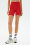 A person wearing SPLITS59 Ella High Waist Airweight Shorts in Pirate Red/Indigo, paired with white socks and white sneakers, standing against a plain white background.