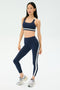A woman wears the SPLITS59 Miles High Waist Rigor 7/8 leggings in Indigo/White with a matching sports bra and silver running shoes, standing against a plain light background.