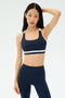 A woman wears the SPLITS59 Miles Rigor Bra in Indigo/White, standing against a plain light background with one hand on her head and a neutral expression.