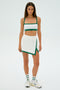 Person in a Monah Rigor Bra - White/Arugula sports outfit by SPLITS59, with a crop top and skirt, white sneakers, and matching socks, standing on a plain white background.