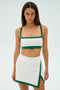 A person wearing the SPLITS59 Monah Rigor Bra in White/Arugula stands against a plain background, dressed in a white and green athletic outfit and skirt.