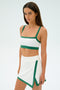 A person with long dark hair is wearing the Monah Rigor Bra in White/Arugula by SPLITS59, paired with a sporty skirt, standing against a plain light background.