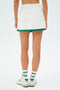 Someone wearing the SPLITS59 Monah Rigor Skort in White/Arugula, paired with white socks featuring green stripes and white sneakers with blue logos, stands against a plain background.