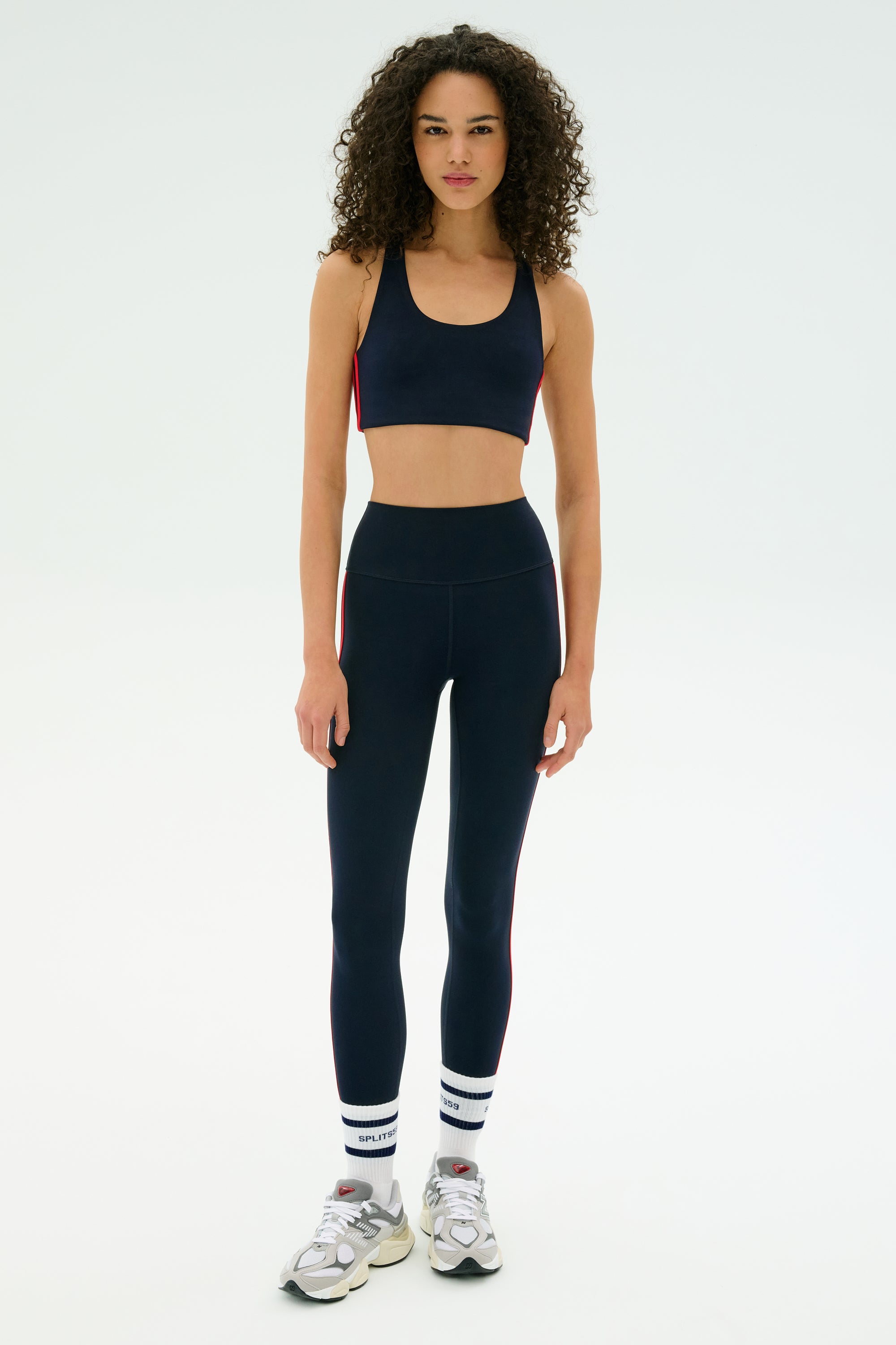 A woman faces forward in SPLITS59's Ella High Waist Airweight 7/8 leggings in Indigo/Pirate Red, styled with a matching sports bra, white striped socks, and gray sneakers—a chic and comfortable look for hot yoga.