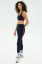 A woman in SPLITS59’s Ella High Waist Airweight 7/8 leggings in Indigo/Pirate Red sports bra, with white socks and silver sneakers, stands against a white background—ideal for hot yoga sessions.