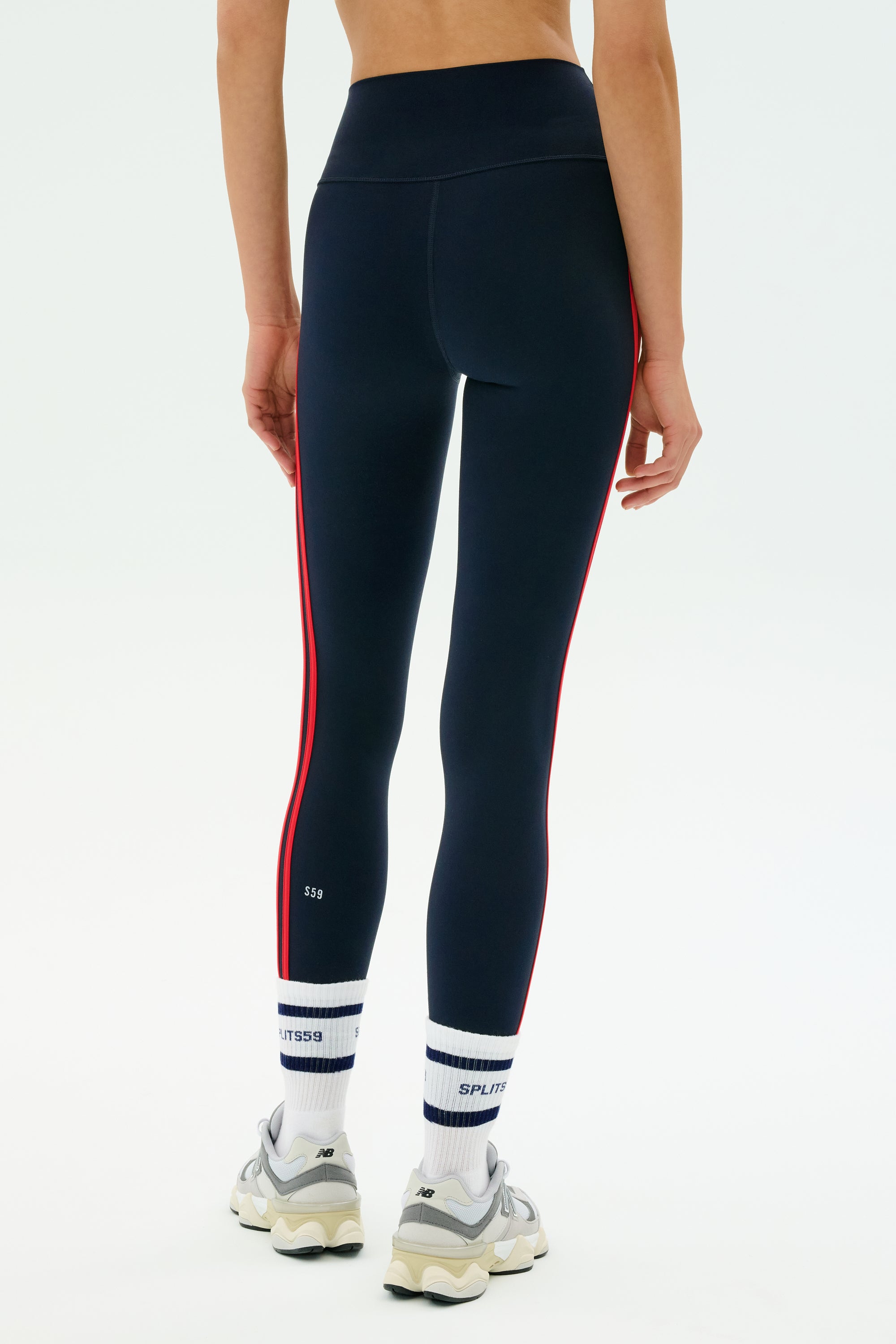 A person faces away, wearing SPLITS59 Ella High Waist Airweight 7/8 black leggings with red side stripes, white socks with blue text, and white athletic shoes against a plain background.