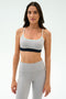 Woman wearing the SPLITS59 Emerson Airweight Bra in Dove Grey/Indigo with thin straps and high-waisted leggings, ideal for hot yoga or Pilates, stands against a plain white background.