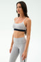 A woman in the SPLITS59 Emerson Airweight Bra in Dove Grey/Indigo and matching high-waisted leggings stands against a white background, ready for hot yoga or Pilates.