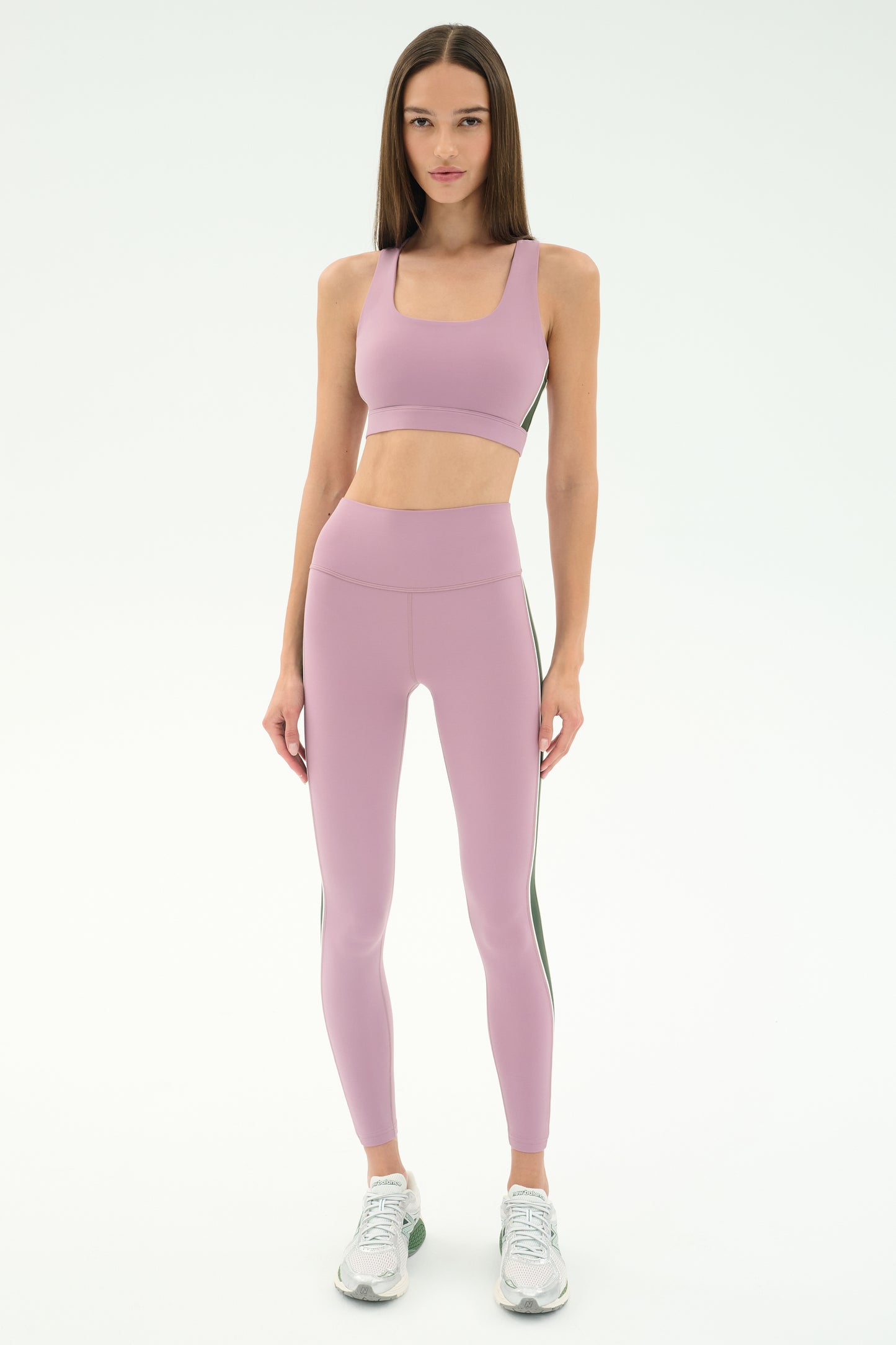 A woman faces forward, wearing the SPLITS59 Lexi Rigor Bra in Blush/Military paired with matching leggings and white athletic shoes, set against a plain light background.