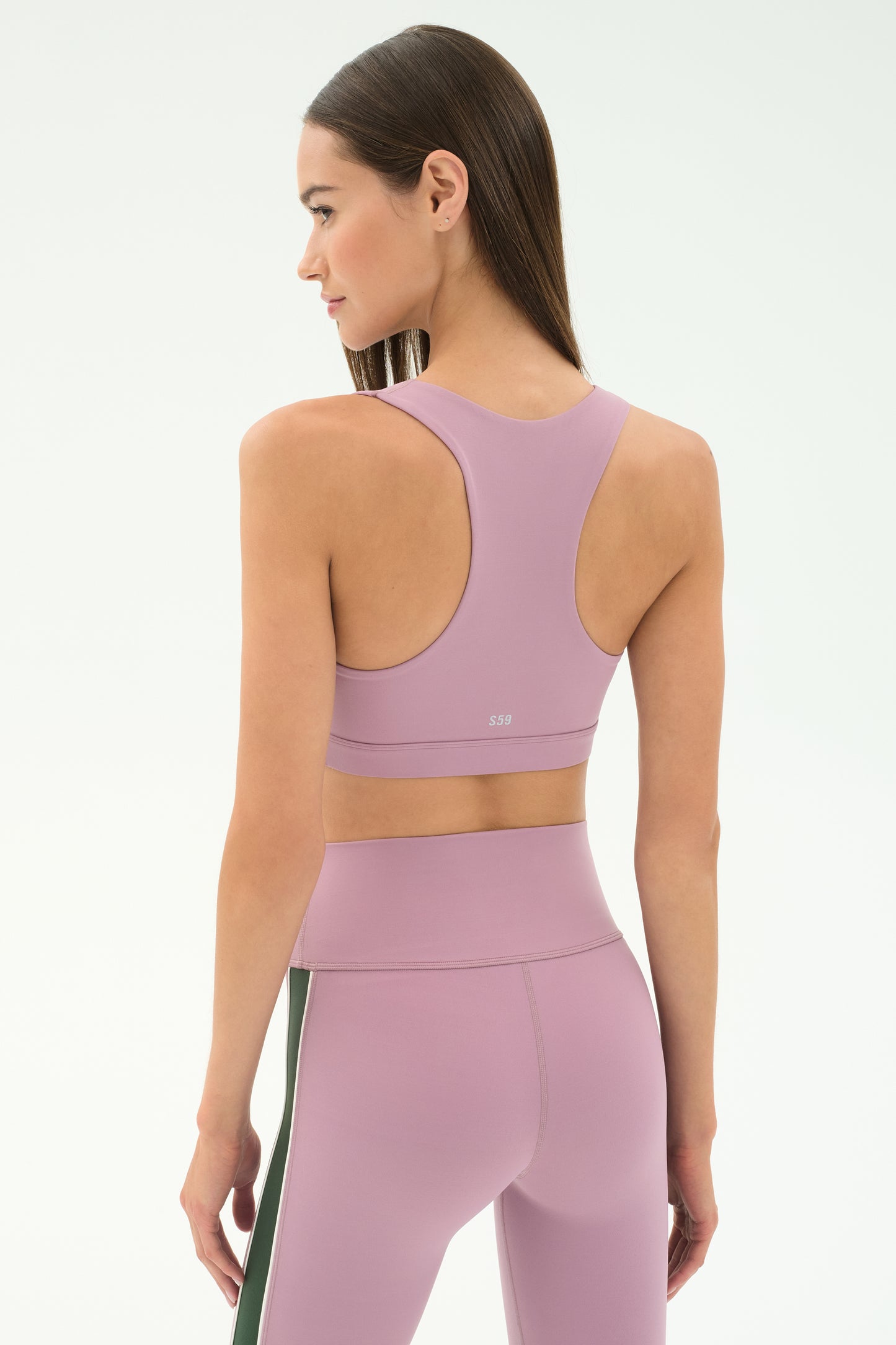 A woman with straight brown hair stands with her back to the camera, modeling the SPLITS59 Lexi Rigor Bra in Blush/Military and matching high-waisted leggings against a plain light background.