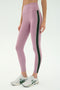 A person wears SPLITS59 Margot Rigor Legging - Blush/Military, high-waisted lavender leggings with a dark green side stripe, paired with white sneakers, standing against a plain light background.