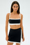 A woman in a SPLITS59 Monah Rigor Bra in black/white and skirt stands against a plain white background, showcasing firm support for intense gym workouts.