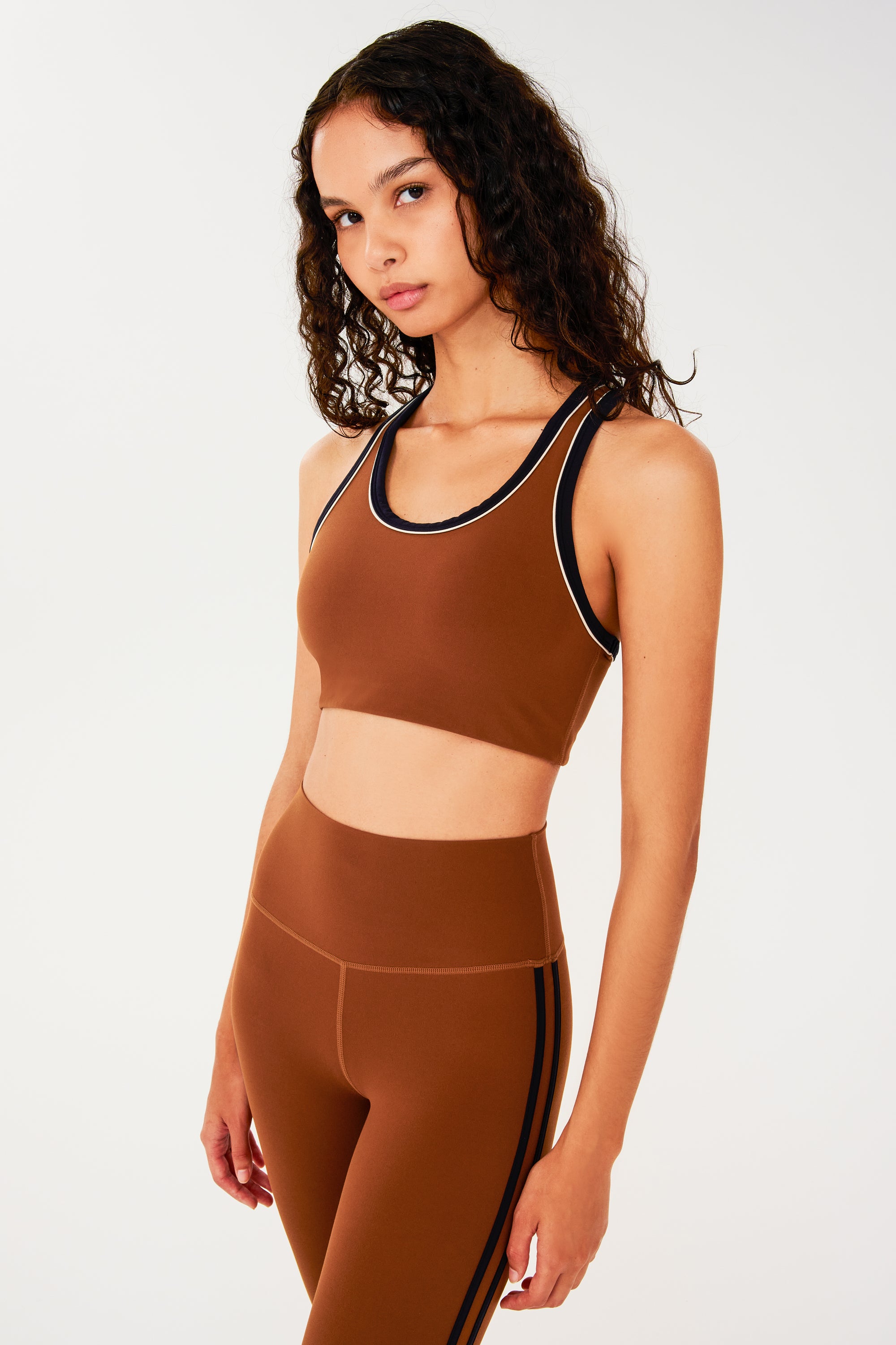 Front side view of woman with dark wavy hair wearing dark orange bra with white and dark blue border arm and neckline and racerback and dark orange leggings with dark blue double side stripes.