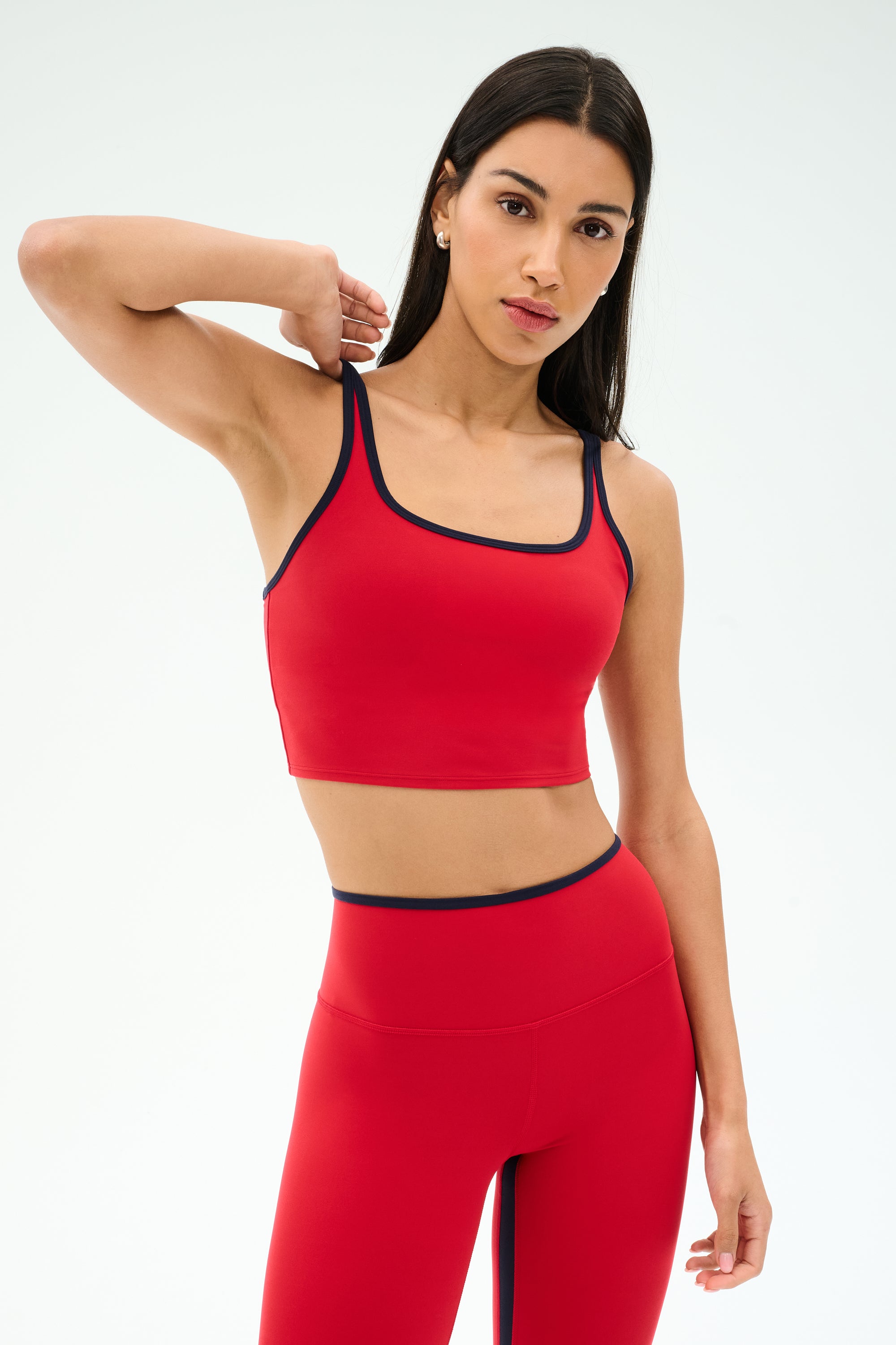 A woman models the SPLITS59 Cait Rigor Bralette in Pirate Red/Indigo, posing with one arm behind her head against a plain white background.