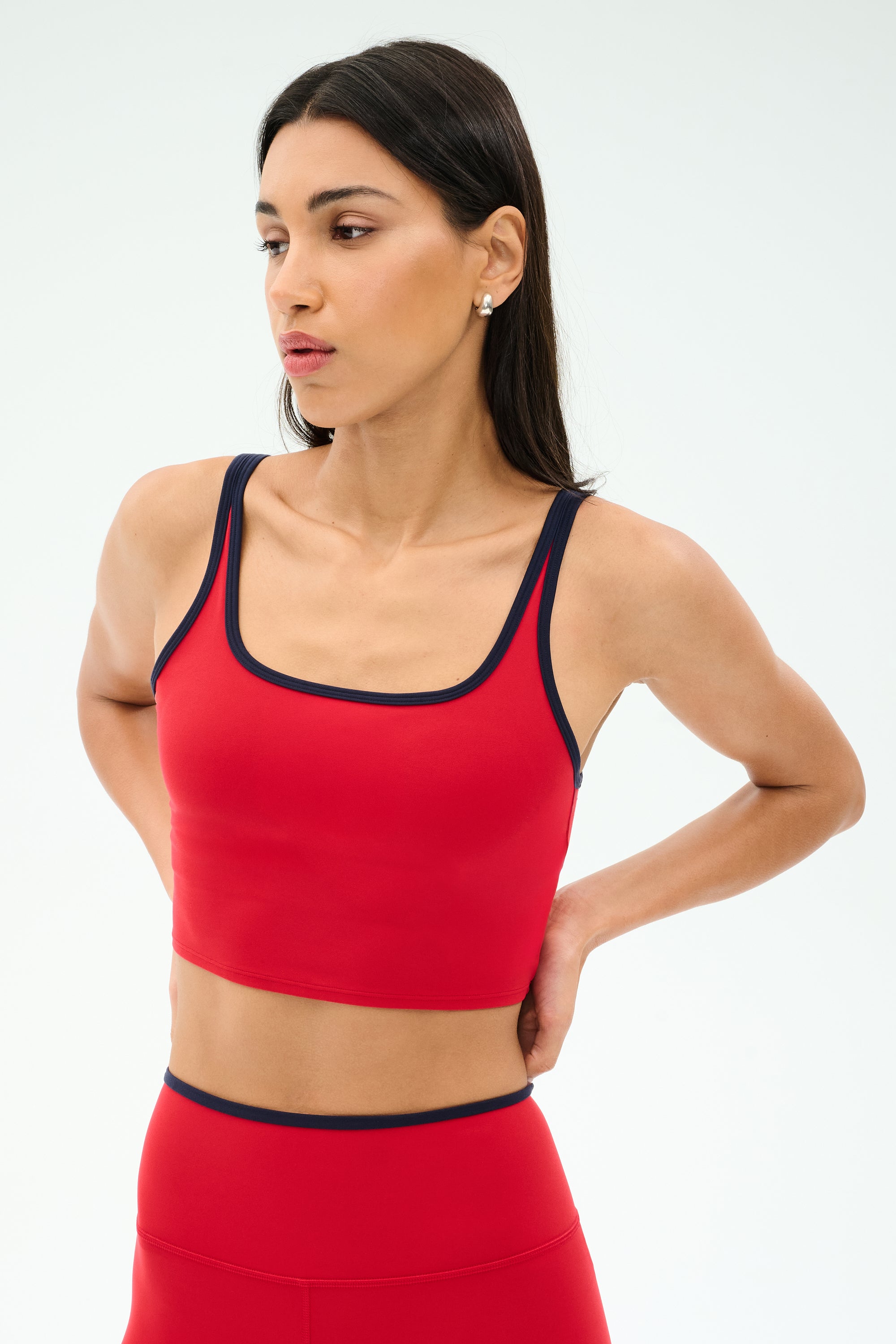 A woman models the SPLITS59 Cait Rigor Bralette in Pirate Red/Indigo, featuring black trim, with matching leggings, standing hands behind her back and looking to the side against a plain background.