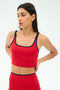 A woman models the SPLITS59 Cait Rigor Bralette in Pirate Red/Indigo, featuring black trim, with matching leggings, standing hands behind her back and looking to the side against a plain background.