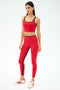 A woman wears the SPLITS59 Cait Rigor Bralette in Pirate Red/Indigo, paired with matching leggings and white athletic shoes with red stripes, standing against a plain white background.