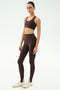 A person stands against a plain background wearing SPLITS59's River High Waist Airweight Stirrup leggings in Dark Chocolate, paired with a matching sports bra and white sneakers.