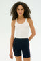 A person with curly hair wears the SPLITS59 Ashby Rib Crop tank in white and navy blue high-waisted shorts with red side stripes, standing against a plain light background.