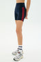 A person wears SPLITS59 Ella High Waist Airweight Short in Indigo/Pirate Red, paired with white socks and gray sneakers, standing against a plain white background.