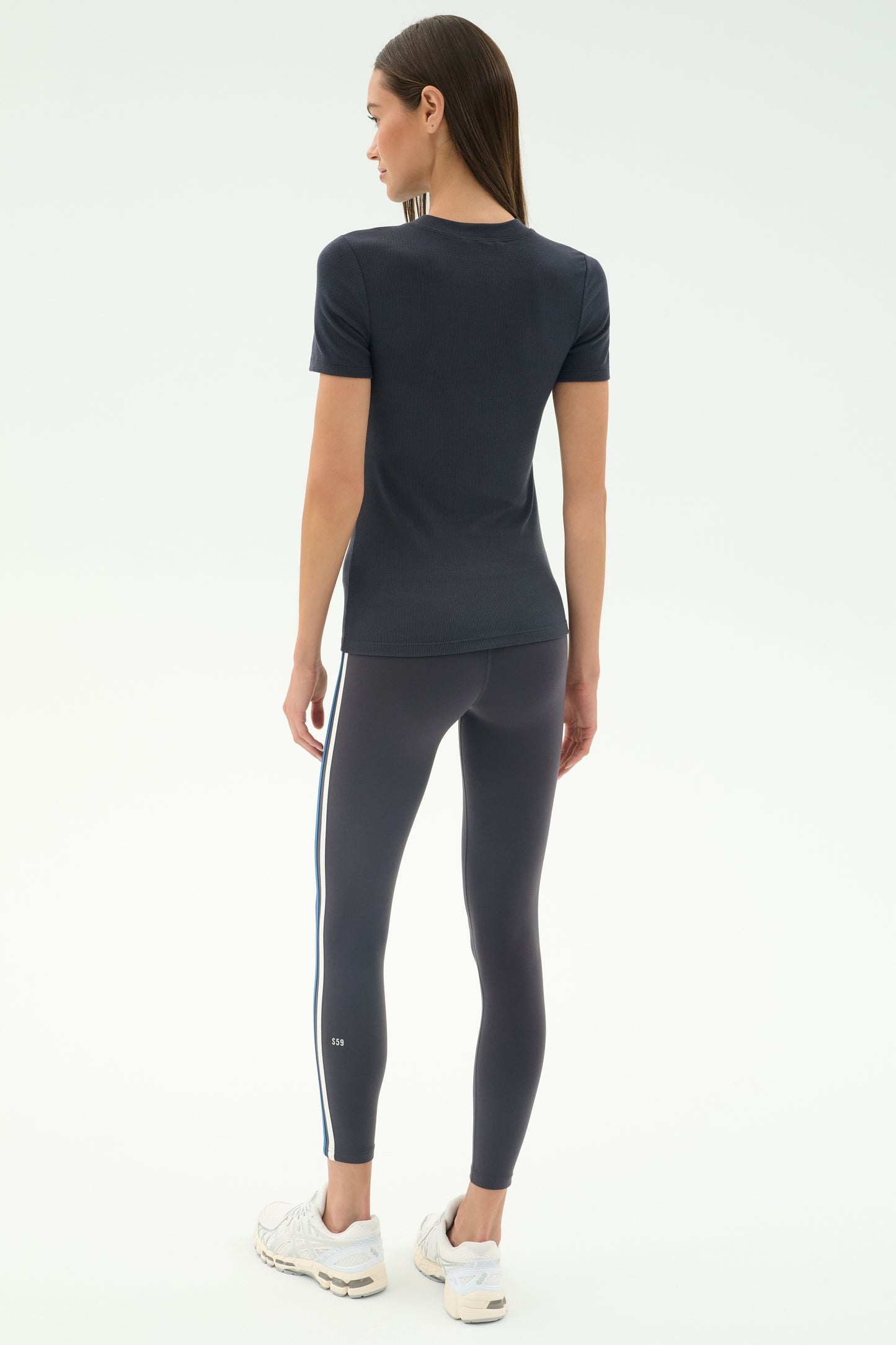 A woman faces away, wearing the SPLITS59 Louise Rib Short Sleeve in Graphite, dark leggings with side stripes, and white sneakers against a plain light background.