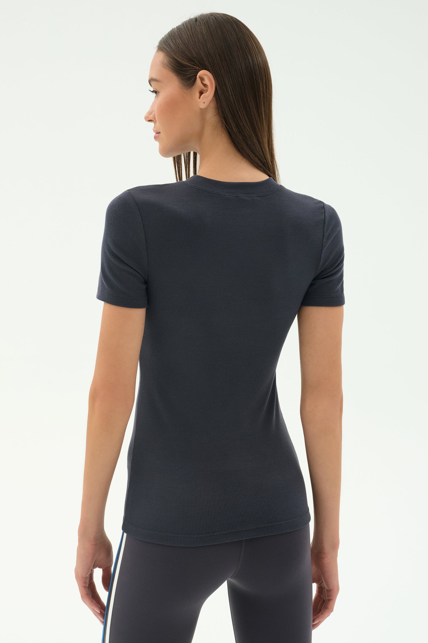 A woman stands with her back to the camera in a fitted SPLITS59 Louise Rib Short Sleeve - Graphite tee and dark leggings, set against a plain background.