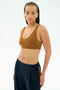 A person wearing the SPLITS59 Airweight Scoop Back Bra in Honey and dark blue jogger pants stands against a plain light background, looking directly at the camera.