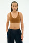 A woman with long braids wears the SPLITS59 Airweight Scoop Back Bra in Honey and dark pants, standing against a plain white background with her hands in her pockets.