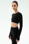 A woman stands against a plain background wearing the SPLITS59 Amelia Airweight Lite Shrug in black, layered over a matching long-sleeve crop top and high-waisted leggings—ideal for yoga or dance warmup sessions.
