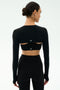A woman in a fitted black long-sleeve crop top with open back cutouts and matching high-waisted leggings, styled with the SPLITS59 Amelia Airweight Lite Shrug in Black, stands against a plain light background.