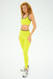 A person with curly hair stands on a plain background, slightly turned to the side, wearing a vibrant chartreuse SPLITS59 Cait Rigor Bra for high-impact workouts, paired with an athletic outfit and sneakers.