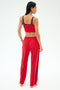 A woman faces away, wearing a red sports bra and SPLITS59 Maxie Rigor Track Pant in Pirate Red/Indigo—wide leg track pants with indigo trim—against a plain light background.