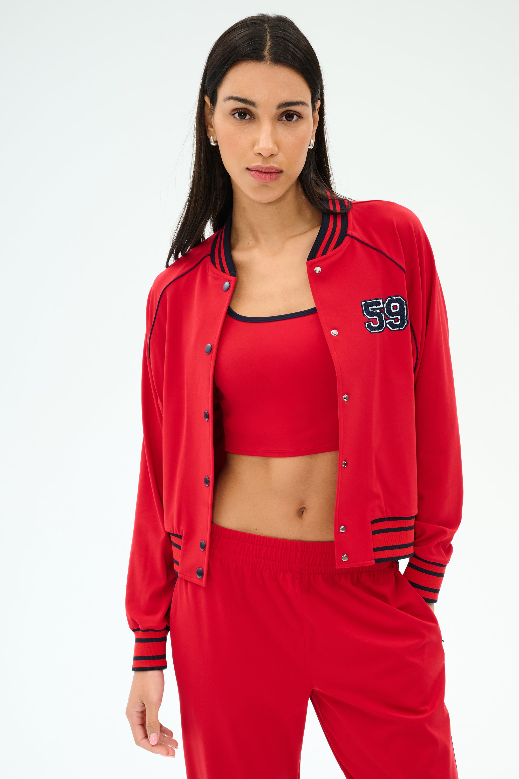 A woman wearing the SPLITS59 Hayes Varsity Rigor Jacket in Pirate Red, paired with a matching red crop top and red nylon spandex pants, stands against a plain light background.