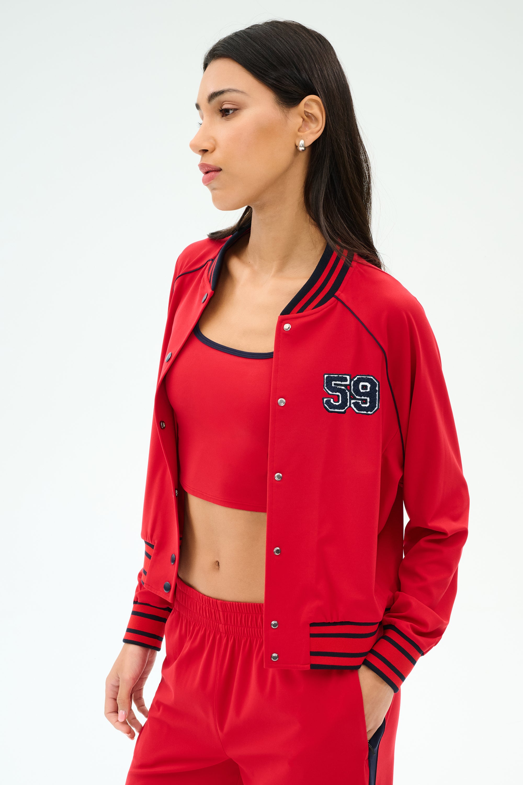 A woman in the SPLITS59 Hayes Varsity Rigor Jacket in Pirate Red, crafted from smooth nylon spandex fabric.