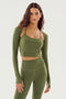 A woman with long hair wears the Shiri Airweight Shrug in Olive, a long-sleeve cut-out crop top by SPLITS59, paired with matching high-waisted leggings made from a nylon-spandex blend. She stands against a plain, light background.