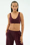 A woman wears the SPLITS59 Airweight Scoop Back Bra in Dark Cherry, made from soft nylon spandex, paired with matching pants and stands against a plain light background with one hand in her pocket.