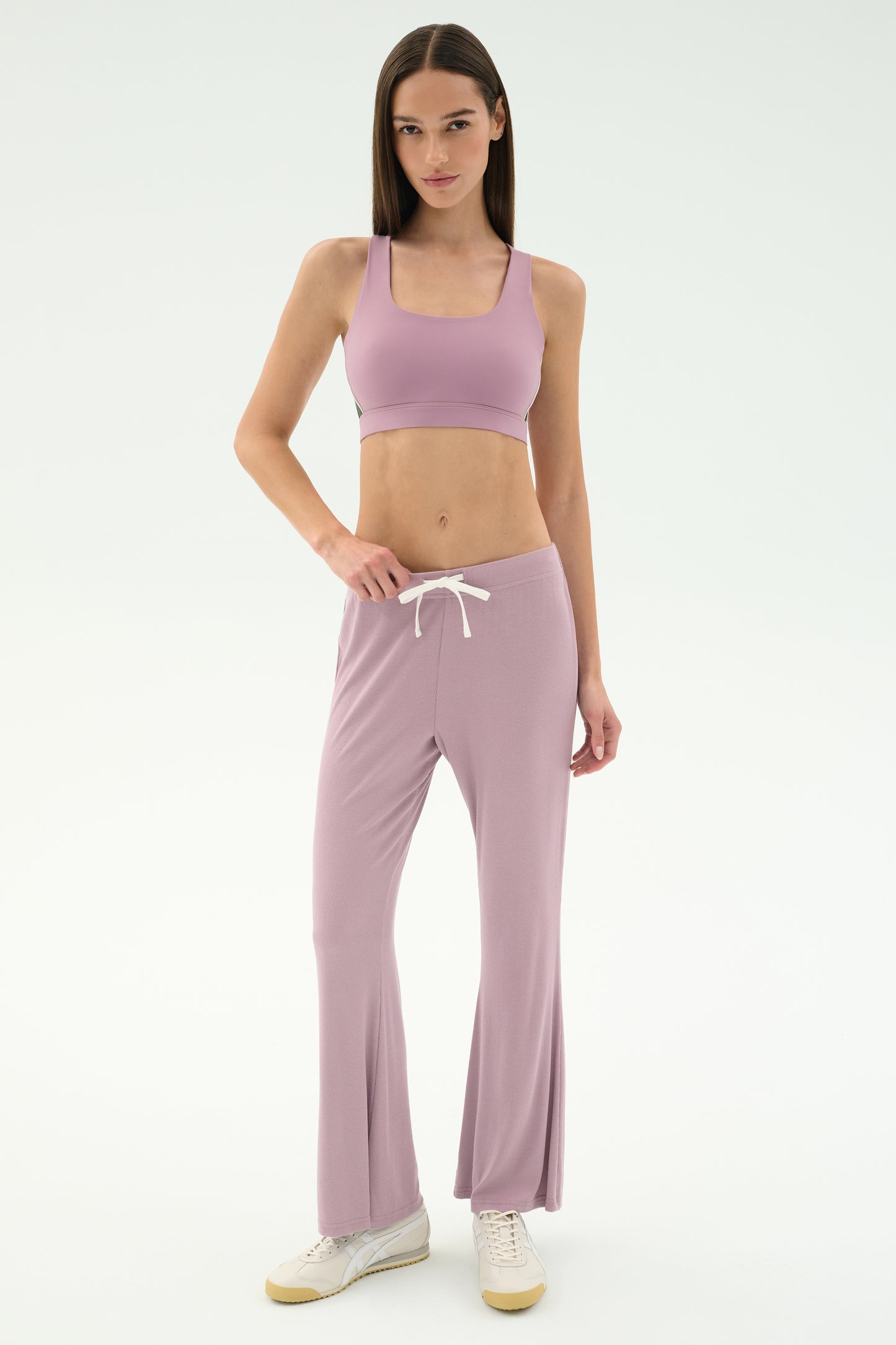 A woman stands against a white background wearing SPLITS59's Blaire Rib Pant in Blush, a coordinating mauve spandex sports bra, and white sneakers.