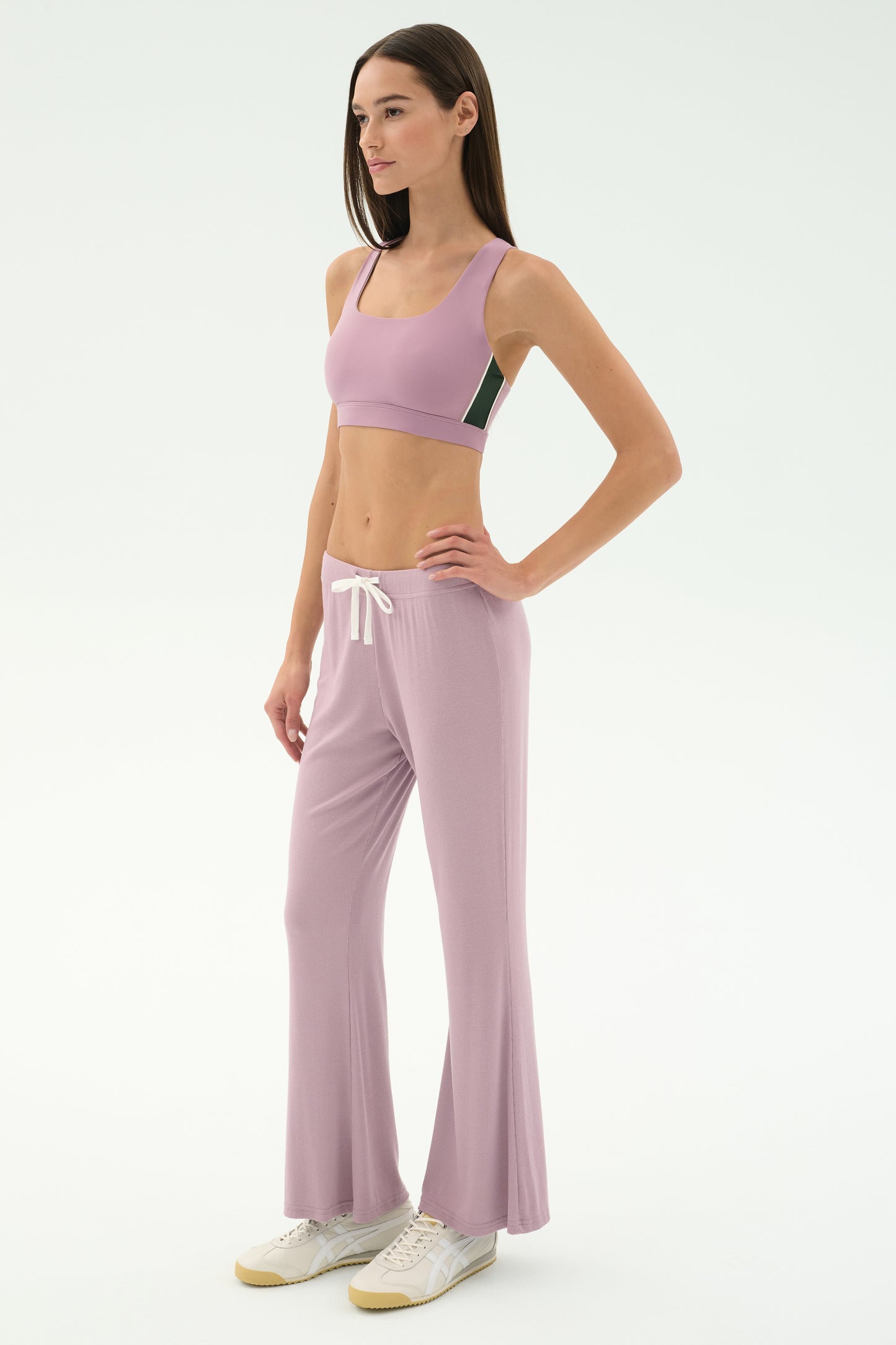 A woman, facing right against a plain white background, wears the SPLITS59 Blaire Rib Pant in Blush with a matching lilac modal-spandex sports bra and white sneakers.
