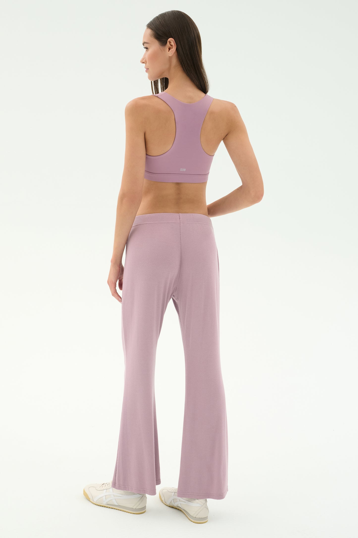 A woman faces away, wearing the SPLITS59 Blaire Rib Pant in blush with a matching lavender sports bra and white sneakers, set against a plain, light background. The outfit is crafted from soft modal fabric with added spandex for comfort.