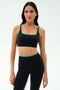 Woman wears the SPLITS59 Cait Rigor Bra in Black/Grass, featuring green trim and high-performance Rigor fabric—ideal for high-impact workouts. She stands confidently against a plain white background.